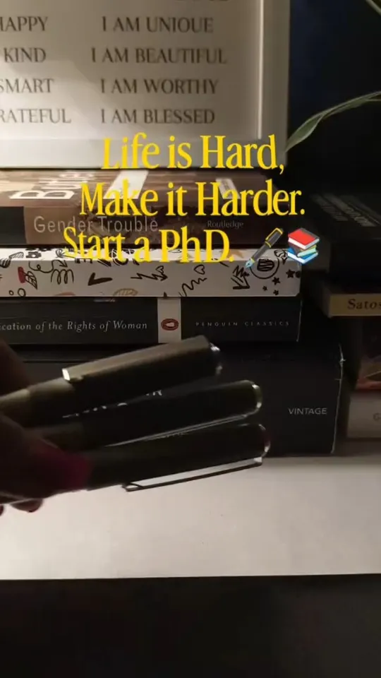 A desk of stacked books and pens below a wall of affirmations, captioned "Life is Hard, Make it Harder. Start a PhD."
